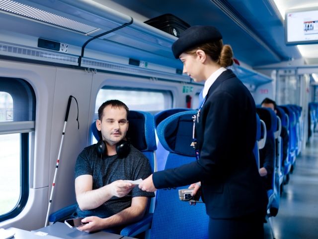 Powiększ zdjęcie: Zdjęcie przedstawia niewidomego mężczyznę, siedzącego w wagonie pociągu i  okazującego bilet konduktorowi. fot. PKP Intercity
